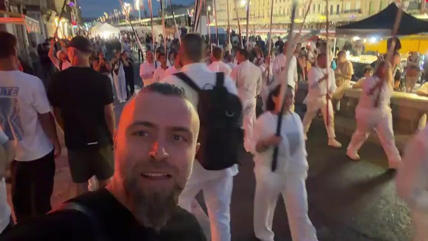 A man with a beard smiles at the camera, holding it up to film a group of people in white outfits. They are marching in a procession, each holding a long white pole. The procession is taking place on a street lined with buildings, possibly at dusk or in the evening, as lights are visible in the background. Other people, dressed in casual clothing, are watching the procession from the side. The man filming is in the foreground, and the procession is in the middle ground and background. The atmosphere appears festive.