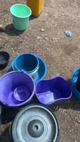 The video shows a group of women standing outside on a sunny day, near several buckets. The buckets are various colors including blue, gray, purple, green and black. Some are stacked. A water hose is visible, and water is running. The women are dressed casually. The background shows a building, a water tower, and greenery. The women appear to be waiting and looking around.