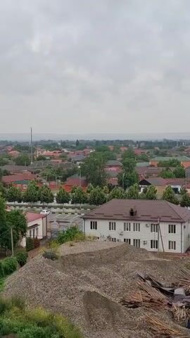 This is a video of a suburban area under a cloudy sky. The camera pans across a landscape of houses with red and brown tiled roofs, interspersed with trees and greenery. In the foreground, a pile of gravel and construction debris is visible. A two-story building with a brown roof is in the lower right corner. The camera pans to the left, showing a parking lot with a white car, and then continues to reveal more houses and trees. The distant horizon is hazy and features rolling hills.