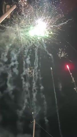 A fireworks display is captured against a dark night sky. Multiple fireworks burst in a variety of colors including pink, green, white, and purple. Some fireworks explode in a large, circular pattern with many individual sparks, while others appear as streaks of light. Wisps of smoke trail from the explosions, creating a smoky atmosphere. Two utility poles with wires are visible in the foreground, one on the left and one on the bottom center of the frame. The overall scene depicts a festive and energetic celebration.