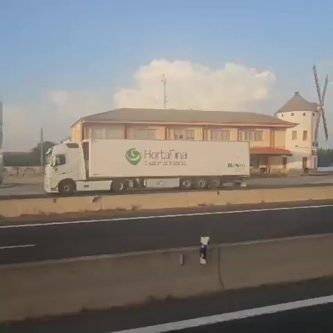 The video shows a highway scene from a moving vehicle. On the left side of the highway, there is a building with a windmill on top. A white truck with "CalaFina" written on its side is driving past the building. The sky is blue with some white clouds. The highway has multiple lanes and is separated by concrete barriers. The right side of the video shows more of the highway, trees, and some signs. The video captures the perspective of someone traveling by car or bus.