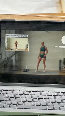 A tablet displays a fitness video. The main screen shows a woman in athletic wear performing an exercise. In the upper left corner, a smaller preview screen shows a woman doing a squat and lunge exercise. The video has a progress bar at the bottom and a timer counting down from 5 seconds. The tablet is placed on a keyboard.