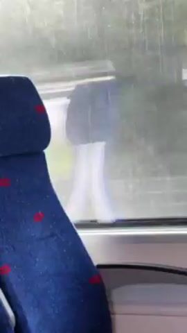 The view from the window of a moving train shows an outside scene, with a blue seat with red markings in the foreground.