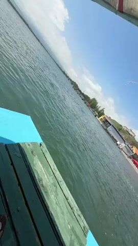 This video captures a view from a wooden pier, angled looking out over a calm body of water. The water is a deep green with subtle ripples, reflecting the bright blue sky. In the distance, a shoreline is visible, featuring a beach with red umbrellas, small buildings, and trees. The scene appears to be a sunny day, possibly late morning or early afternoon, given the strong sunlight and clear skies with a few scattered clouds.

The pier itself has a weathered green wooden surface with a bright blue section at the edge, suggesting it might be part of a dock or a boat. There are no people clearly visible in the immediate foreground, but the presence of beach umbrellas and buildings on the shore indicates a populated area, likely a recreational spot. The landscape slopes upwards behind the beach, with trees and some structures dotting the hillside. The overall atmosphere is peaceful and serene, typical of a leisurely day by the water. The camera angle is tilted, giving a dynamic perspective of the scene.