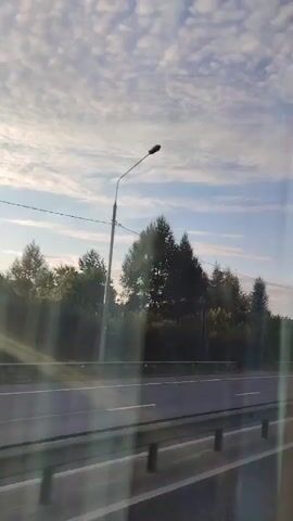 The video is a first-person perspective of a drive along a road. The time is unclear. The sky is visible, with scattered clouds. Trees and buildings are seen on the right side of the road. A street light is visible. There are cars on the road.