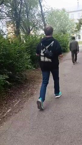 A person on a scooter is moving along a path, and another person is walking ahead of them. The path appears to be made of asphalt and is surrounded by green bushes and trees. In the background, there is a building. The person on the scooter is wearing a black hoodie and blue jeans. The other person is wearing a brown jacket. The lighting suggests it is daytime.