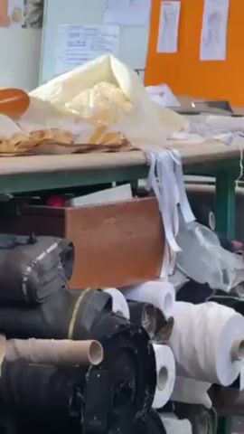 The video shows a sewing or design studio. There are rolls of fabric of various colors and textures piled up, along with boxes and scraps of material beneath a large table. On the table are more piles of fabric, sketches, and a wooden box filled with colorful ribbons. A person wearing grey leggings and a long-sleeved shirt appears and unfolds a large piece of white, transparent fabric on the table. Next to the table is a dress form. The scene suggests a space dedicated to fashion design and creation.