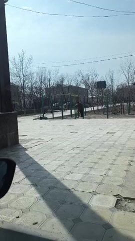 The video depicts an outdoor setting, likely a park or recreational area, on a bright day with a clear, pale blue sky. The ground is paved with geometric, gray tiles that show signs of wear and tear with cracks and discolored areas. To the left of the frame, the edge of a building or wall is visible, with its dark gray surface casting a shadow on the paved ground. A portion of a car mirror is also noticeable.

In the center of the frame, there is playground equipment consisting of green support beams and yellow horizontal bars. Two individuals are standing near the equipment. They appear to be engaged in a conversation or interaction. The figures are dressed in what appear to be casual clothes.

Beyond the playground equipment, several bare trees are scattered throughout the area, suggesting it may be late fall or early spring. Buildings can be seen further in the background, indicating an urban or suburban setting. A basketball hoop and backboard are visible to the right, standing as another element of the recreational area.