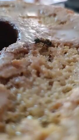A close-up, slightly blurry shot shows a bee on a baked good, possibly a cake or bundt cake, with a shiny glaze. The bee is positioned on the textured surface of the cake, near a darker hole in the center. The bee's body is striped black and yellow. The cake appears to be light brown with some embedded ingredients, and a white, liquid-like glaze is visible on its surface. The background is out of focus, with hints of other objects and a light-colored surface.