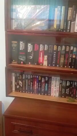 A video shows a bookshelf filled with books. The bookshelf has three shelves, and each shelf is packed with books. The books are of various sizes and colors, and many of them are by the author Stephen King. There are also some other authors' books visible. The bookshelf itself appears to be made of wood. The lighting in the video is somewhat dim, and the camera is shaky, making it difficult to read all of the book titles clearly. The video appears to be filmed in a domestic setting, possibly a living room or study. There are no people visible in the video. The video is very short, lasting only a few seconds.