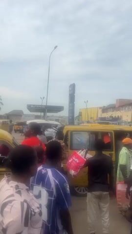 The video shows a busy street scene in what appears to be Nigeria. Several yellow minibuses are parked near each other, with a crowd of people standing around. Some of the people appear to be boarding or exiting the vehicles. In the background, there is an ENYO gas station with visible price signs. The sky is bright but overcast. The video appears to be taken from within the crowd.