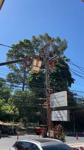A utility worker in an orange shirt and white hard hat is performing maintenance on a utility pole. The worker is attached to the pole by a harness and rope, and is holding a tool to adjust wires. Another utility worker is visible in a bucket truck, also working on the pole. The pole is covered in electrical wires and insulators, and is situated in an outdoor environment with trees and a blue sky. Two signs are attached to the pole, one with text and a QR code, and another with text that appears to read "Be Sweet". A motorcycle and a car are visible in the background.