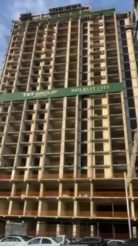 The video shows two high-rise buildings under construction. The buildings are primarily concrete with a grid-like structure and lack exterior walls. A green banner with the words "TXT GROUP" and "BRILIANT CITY" is attached to one of the buildings. A crane is positioned between the two buildings, with its arm stretching across the sky. Cars are parked along the street in front of the buildings. The sky is partly cloudy.
