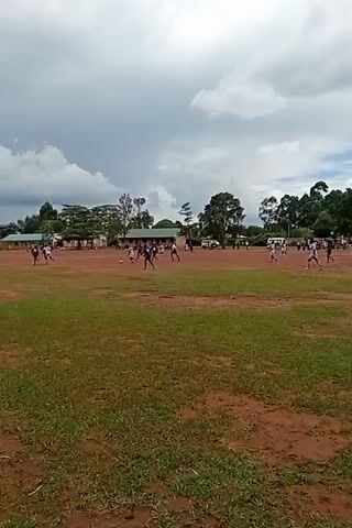In a medium shot on a cloudy day, people are playing on a dirt field. There are 26 people in total, all wearing shorts and t-shirts. The t-shirts are black or white, the shorts are black, white, or blue. The field is mostly dirt with some patches of green grass. Behind the players are buildings and trees. In the background are vehicles parked behind the buildings. The camera is stationary.