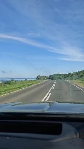 The view from the dashboard of a moving vehicle shows a two-lane road with double white lines running down the center, leading towards the horizon. To the left of the road is a body of water, possibly a lake or bay, with a visible shoreline. The sky above is blue with streaky white clouds. On the right side of the road, there is green vegetation and trees, which rise slightly to form a small hill. Power lines are visible along the roadside. The day appears to be sunny.