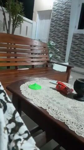 This video captures a close-up view of a wooden table in an outdoor setting, likely a patio or porch, at night. The table is covered with a white lace doily. On the doily, there are a few items: a bright green circular object that appears to be a toy or a lid, a red and silver object that looks like a pack of cigarettes or candy, and a dark, possibly ceramic, pot or ashtray. To the left of the table, a portion of a wooden bench is visible, with a person's patterned clothing on the very left edge of the frame, suggesting someone is sitting there.

In the background, a wooden bench is positioned against a wall with a patterned texture. There's also a white door and some greenery, including a tree with sparse leaves. The lighting suggests it's nighttime, with artificial light illuminating the scene. The overall atmosphere seems calm and relaxed, potentially indicating a casual evening gathering or a moment of quiet observation. The location context of Panjalin Kidul, Indonesia, implies a warm, tropical climate, though no specific weather details are discernible from the video itself. No visible text is present in the frame.