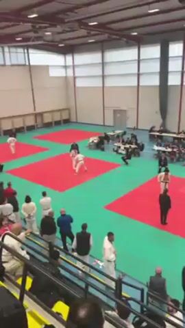 The video shows a judo match in progress. Two men in white judogi are competing on a red and green mat. One has a purple belt and the other a black belt. An older man in a suit observes as the referee. The mats are surrounded by spectators in a gymnasium. Some of the spectators are standing near the edge of the mat, while others are seated on tiered seating. The gymnasium has high ceilings with skylights.
