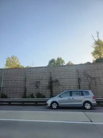 This video captures a view from a moving vehicle alongside a highway. The primary focus is a sound barrier wall, constructed from rectangular concrete panels with a textured surface. The wall is bordered by a metal guardrail at its base. Beyond the wall, a clear blue sky dominates the upper portion of the frame, with glimpses of trees and foliage peeking over the top of the barrier in some segments. In one section of the video, transparent panels are interspersed with the concrete, reflecting the surrounding environment. The scene appears to be a rural or suburban setting, devoid of any visible people or specific identifiable landmarks. The lighting suggests it is daytime, with the sun casting some glare in certain shots.