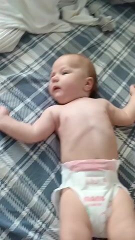 This is a short video clip of a baby lying on its back on a bed with a plaid comforter. The baby is wearing a diaper. The baby's arms are extended to its sides. The baby's head is turned to the left, and it appears to be looking at something off-camera. The baby's mouth is slightly open, and it may be speaking or making a sound. The lighting suggests it is daytime, but no other weather or specific location cues are visible. The overall mood is calm and relaxed. The diaper has some text and graphics on it, including the word "COMFORT" in pink lettering and a cartoon design. The baby appears to be content and alert.