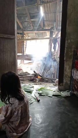 A girl wearing a pink and white patterned pajama set sits on the floor with her back to the camera. She is facing an open doorway that looks out onto an outdoor area where a fire is burning. Banana leaves are spread on the floor around the girl. To the right of the doorway, a person wearing a dark blue dress stands facing away from the camera, looking into the outdoor area. Smoke is rising from the fire. The indoor space appears to be a home with a dark floor and textured walls.
