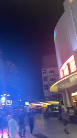 This media file captures a lively street scene at night in Patong, Thailand. The overall impression is one of vibrant nightlife and bustling activity.

The primary subjects are people walking along a paved walkway. Silhouettes and dimly lit figures suggest a crowd moving through the area. In the foreground, several individuals are visible, including a person in a light-colored outfit and another in darker clothing. Further back, more people can be seen in groups and individually.

The setting is an urban street illuminated by a mix of neon lights, streetlights, and spotlights. Tall palm trees punctuate the scene, their fronds silhouetted against the dark night sky. Buildings with multiple stories line the street, their facades adorned with signs and illuminated windows. Several vehicles are present, including yellow minibuses that appear to be taxis, and other red vehicles, possibly tuk-tuks or similar transport. The bright blue and pink lighting cast a dynamic glow over the environment.

The activity taking place is clearly people enjoying an evening out. The presence of illuminated establishments, the movement of people, and the general atmosphere suggest a commercial and entertainment district. The time of day is unequivocally night, indicated by the dark sky and artificial lighting. There are no explicit weather cues, but the lighting suggests it is likely dry.

Notable details include the striking use of colored lights, which create a visually stimulating and somewhat hazy effect. The signage on the buildings is partially visible, with some text legible. One sign reads "HOLLYWOOD" in large, illuminated letters. Another sign, higher up and further back, appears to have "LIVE" on it. Smaller signs with text and images are scattered throughout the background, indicating various businesses or attractions. The overall emotion conveyed is one of energy and a lively atmosphere, typical of a popular tourist destination at night.