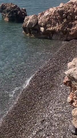 The video shows a view of a rocky beach and the sea. The water is clear and calm, with small waves lapping at the shore. There are several large rocks in the water, and more rocks are visible on the beach. The sun is shining, creating a bright and serene atmosphere. The footage appears to be filmed from a stationary position on the beach, looking out towards the sea and rocks. There are no people visible in the video.