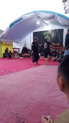 The video shows a performance outdoors under a tent. Two individuals in dark clothing are engaged in what appears to be a martial arts demonstration on a red carpeted stage. One performer is in the foreground, facing away from the camera, while the other is further back and to the left. Several onlookers are seated behind the performers. A banner with text and a logo is visible on a black backdrop behind the stage. Musical instruments, including drums, are present on the left side of the stage. The overall environment appears to be a rural setting with some buildings visible in the background.