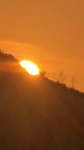 The video shows a sunrise over a hilly landscape. The sun is partially visible, casting a warm orange glow across the sky. The silhouette of a hill with sparse vegetation is visible in the foreground. The sun is positioned on the left side of the frame, peeking over the crest of the hill. Rays of sunlight emanate from the sun, creating a dramatic effect. The overall mood is serene and peaceful.