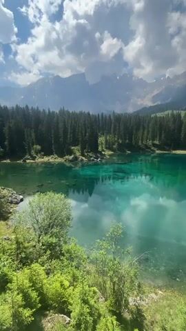 This is a vertical video of a scenic lake surrounded by trees and mountains. The lake has vibrant turquoise water that reflects the sky and the surrounding landscape. The sky is blue with fluffy white clouds. The foreground is filled with lush green foliage. The video appears to be a slow pan across the lake and the scenery. There are no people or specific actions visible.