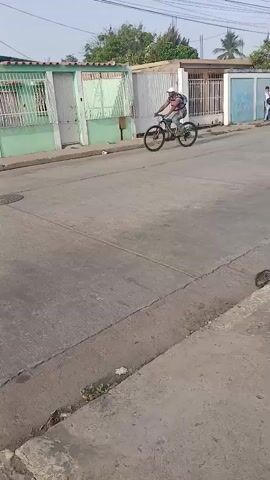 A man rides a black bicycle down a concrete road. He wears a white baseball cap, a maroon shirt, blue jeans, and a blue backpack. The road runs parallel to a row of houses with green and white barred gates. The sky is overcast. Electrical wires span the road above.