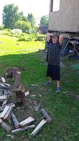A young boy is chopping wood in a rural setting. He is wearing a dark t-shirt with a pattern and dark shorts, and blue sandals. He is standing in a grassy area, with a pile of chopped wood in front of him. The boy is in the process of swinging an axe to chop a log that is resting on a tree stump. He is looking at the log intently, and his mouth is open as if he is exerting effort. The scene appears to be taking place outdoors during the daytime, with sunlight casting shadows on the ground. In the background, there are trees, a greenhouse, and a partially constructed building. The overall atmosphere seems to be one of a typical summer day in a rural area. There is no visible text in the media file.