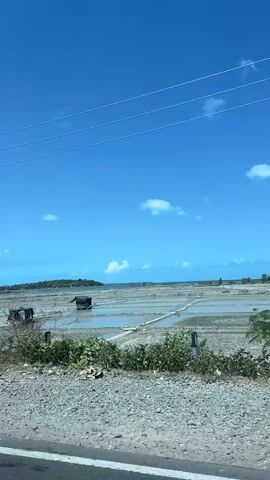 The video shows a view from a moving vehicle on a sunny day with a clear blue sky and a few scattered clouds. In the foreground, a paved road is visible with a white line marking the edge. Beyond the road, there is a dry, gravelly area with sparse vegetation. Further back, a wide expanse of what appears to be salt pans stretches across the landscape, with some areas filled with water. There are small structures or shacks scattered within the salt pans, and some lines of debris or materials are visible. In the distance, there is a strip of land with trees and vegetation, and a body of water. Power lines run across the sky. The camera pans slightly to the right, revealing more of the salt pans and eventually a building with a corrugated metal roof and a large, slatted gate.