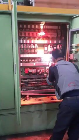A person wearing a blue shirt, a gray safety vest, and dark pants is working on an open electrical control panel. The panel is filled with numerous components, including relays, circuit breakers, and wiring. A red light is illuminated within the panel, casting a warm glow on the scene. The person is holding a tool and appears to be making adjustments to the wiring or components. The environment appears to be an industrial setting, with hints of machinery in the background.