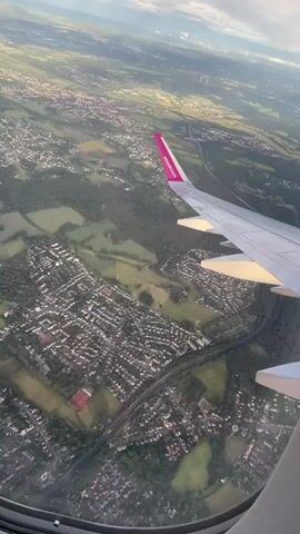 The video captures an aerial perspective from an airplane window, looking down upon a diverse landscape beneath a partly cloudy sky. Dominating the foreground is the right wing of the aircraft, extending from the bottom right towards the center of the frame. Its distinctive wingtip is painted a bright magenta, emblazoned with the white text "wizzair.com." The wing itself is illuminated by a warm, golden sunlight, suggesting either late afternoon or early evening.

Below, the terrain is a mosaic of suburban development, green agricultural fields, and dense patches of woodland. Numerous residential houses with varying roof colors, predominantly gray, are clustered together, interconnected by a network of roads. A prominent, dark-colored highway or major road cuts through the scene, winding between the settlements and open fields. In some areas, power lines or utility poles are visible.

The sky shows a mix of white clouds and clear blue patches, with good visibility extending to the horizon where faint structures, possibly wind turbines, can be discerned. The aircraft appears to be in a gentle bank, indicative of either ascent after takeoff or approach for landing. The scene offers a characteristic view of the European countryside and residential areas, consistent with the provided location context of Holzwickede, Germany.