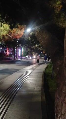 The video is taken at night on a sidewalk next to a road. The sidewalk has a tactile paving system with raised parallel lines. Several people are walking on the sidewalk. A motorcycle passes by on the road, followed by a car. Trees line the sidewalk, and lights are visible in the background, suggesting buildings or businesses.