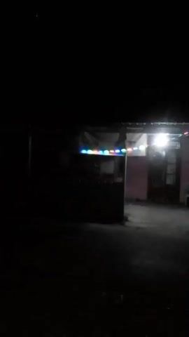 The video shows a nighttime scene outside a small building. The building appears to be a house or a shop, with a pink facade and a covered entrance. There are string lights with colorful bulbs illuminating the front of the building. Inside the building, there are windows and a door. A white bench is visible outside the building, and a vehicle, possibly a car or a van, is parked to the right. The ground in front of the building is unpaved and appears to be dirt or gravel. The overall impression is of a simple, possibly rural, setting at night, with some decorative lighting.