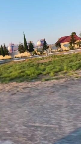 THE VIDEO SHOWS AN EXTERIOR VIEW FROM A MOVING VEHICLE. THE SCENE APPEARS TO BE A SEMI-RURAL AREA WITH SOME INDUSTRIAL STRUCTURES VISIBLE IN THE DISTANCE. THE FOREGROUND IS DOMINATED BY A GRAVEL OR DIRT ROAD. TO THE RIGHT, THERE IS A SHADOW CASTING ACROSS THE ROAD, SUGGESTING THE SUN IS TO THE LEFT AND BEHIND THE VIEWER. IN THE MID-GROUND, THERE ARE AREAS OF GRASS AND SCRUB VEGETATION. FURTHER BACK, THERE ARE SEVERAL TALL, SLENDER TREES AND SOME LARGER, BUSHIER TREES. IN THE FAR BACKGROUND, SEVERAL INDUSTRIAL BUILDINGS WITH TANKS AND PIPING ARE VISIBLE, ALONG WITH A GAS STATION UNDER A BLUE CANOPY. THE SKY IS CLEAR AND BLUE, INDICATING DAYTIME. THE OVERALL IMPRESSION IS OF A DRIVE ALONG A ROAD PASSING BY OPEN LAND AND SOME INFRASTRUCTURE.