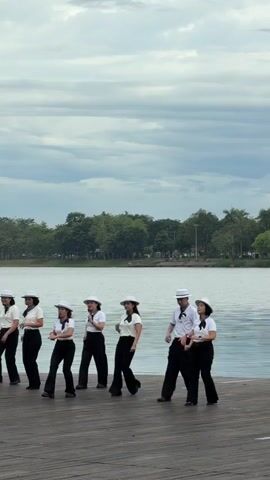 A group of eight people, seven women and one man, are dancing on a wooden pier. They are all wearing white shirts, black pants, and white hats. The women have black scarves around their necks. They are positioned in a line, with the man at the right end of the line. They appear to be performing a choreographed dance, with their bodies angled in various directions. The background shows a large body of water, trees, and a cloudy sky. The lighting is overcast.