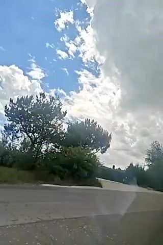 The video shows a view from a vehicle moving along a paved road, with trees and vegetation on the right side and a cloudy blue sky above. The road appears to curve to the right. The vegetation includes trees with dark green foliage and some lighter green bushes. In the distance, there are hints of buildings or structures visible through the trees. The clouds are white and voluminous, suggesting a partly cloudy day. The perspective indicates the video is captured from inside a moving car, likely from the windshield or side window.