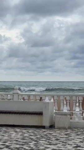 A view of a cloudy sky over a turbulent sea. In the foreground, a white ornate railing separates the viewer from the water. A concrete bench with a wooden seat is positioned in front of the railing. The ground is paved with a black and white checkered pattern. The video shows the waves moving in the sea and the clouds shifting in the sky.