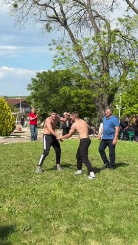 Two shirtless men in athletic pants engage in wrestling on a grassy field, supervised by a man in a blue collared shirt.  Spectators stand around the perimeter of the field. There is a building with an orange tiled roof in the background, along with trees, bushes, and a utility pole. The sky is partially cloudy.