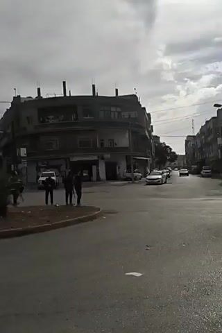 The video depicts a street scene in what appears to be a Middle Eastern city. The sky is overcast. Several multi-story buildings are visible, one with a rounded corner facing the intersection. Cars are parked along the streets and driving through the intersection. There are a few people standing on the sidewalk near the corner. A red pickup truck drives along the road.