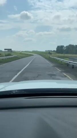 The video shows a view from the inside of a car driving on a paved road. The road is surrounded by green fields with some trees in the distance. The sky is blue with scattered white clouds. The car is moving forward, and the road ahead curves to the left. There are white lane markings on the road, and a guardrail is visible on the right side. A white car is visible in the distance on the left side of the road. The dashboard of the car is visible in the foreground.
