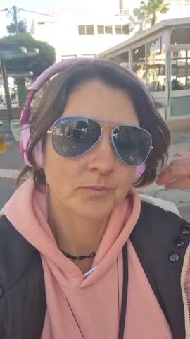 A woman is the primary focus of the video. She is wearing pink headphones, a pink hoodie, a black jacket, and blue aviator sunglasses. She has glitter on her face and is standing outside in a sunny location. She is holding her sunglasses and looks at the camera, removing and adjusting her sunglasses. Behind her, there is a building and a palm tree. The background suggests an urban or coastal environment.