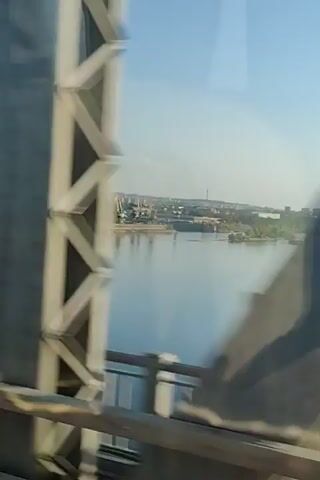 The video captures a view from a bridge overlooking a wide, calm body of water, likely a river. The water is a muted blue, reflecting the clear, bright sky above. On the far bank, a city skyline is visible, with a mix of low-rise buildings and some taller structures, possibly industrial cranes or towers. There are barges or boats floating on the water. The bridge railing is in the foreground, with its metal bars and wooden or concrete supports. As the video progresses, the perspective shifts slightly, showing different segments of the bridge's structure, including a large concrete arch that partially obstructs the view at one point. The sun appears to be shining, creating a bright, daylight scene. There are no people or animals visible in the video.