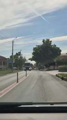 The video shows a view from inside a vehicle driving down a road. In the foreground, the dashboard and windshield are visible. The road is asphalt with white lane markings and a red stripe on the side. On the left side of the road, there are houses, trees, and utility poles with overhead wires. On the right side, there are trees and a sidewalk. Several vehicles are on the road, including a large white truck with a cylindrical tank, a black van, and other cars. The sky is blue with scattered white clouds and contrails. The overall scene depicts a typical daytime scene on a road in a suburban or rural area.