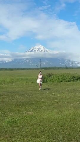 A woman with dark hair, wearing sunglasses, a white crop top, and a white skirt, runs across a grassy field towards the camera. In the background, a snow-capped mountain is partially obscured by clouds. The sky is blue with white clouds.