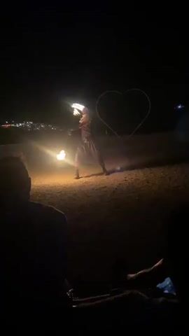 A fire performer in dark clothing, possibly a costume, is spinning a flaming object in a large open area at night. The performer is in the center of the frame, illuminated by the fire and a light source attached to the spinning object. Behind the performer, a large, outlined heart shape is visible, also illuminated by the performer's lights. The background shows a dark sky with distant lights, suggesting a town or city. In the foreground, the silhouettes of several people are visible, appearing to be an audience. The ground is rough and appears to be dirt or sand. The flames are bright and dynamic, creating light and shadow play. The performer's movements are energetic and controlled as they manipulate the fire. The overall atmosphere is one of a nighttime outdoor performance.