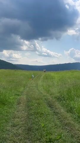 The video shows a wide, grassy field with a dirt path leading up a gentle slope. The sky is partly cloudy with dramatic dark clouds on the left and brighter clouds on the right. In the foreground, a young child in a white shirt and hat is walking on the path, holding an orange object. Further up the path, another person, also wearing a hat, is walking away from the camera. In the background, there is a line of trees and hills. The overall impression is of an outdoor recreational activity in a natural setting.