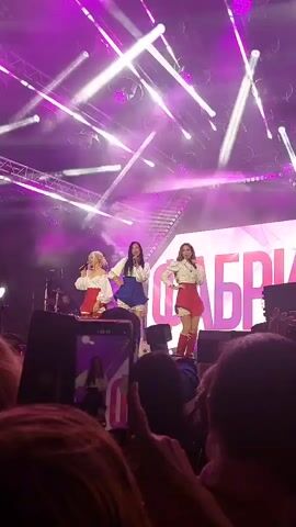 A video shows a concert with three women performing on stage. They are dressed in matching outfits consisting of white blouses with blue trim and red skirts. They are also wearing tall red boots. The stage is lit with purple and white lights, and there are large speakers in the foreground. The audience is visible in the foreground, with some people holding up their phones to record the performance. The word "Фабрика" (Fabrika) is visible in large letters on the stage behind the performers. One of the performers is holding a red object.